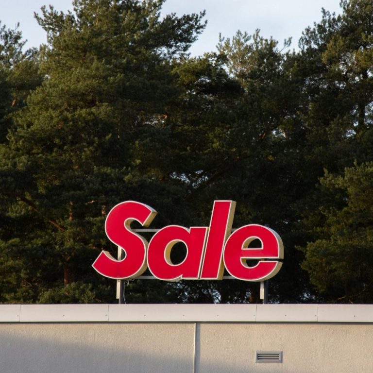Bright red "Sale" sign on top of a building and against a backdrop of trees and cloudy sky.